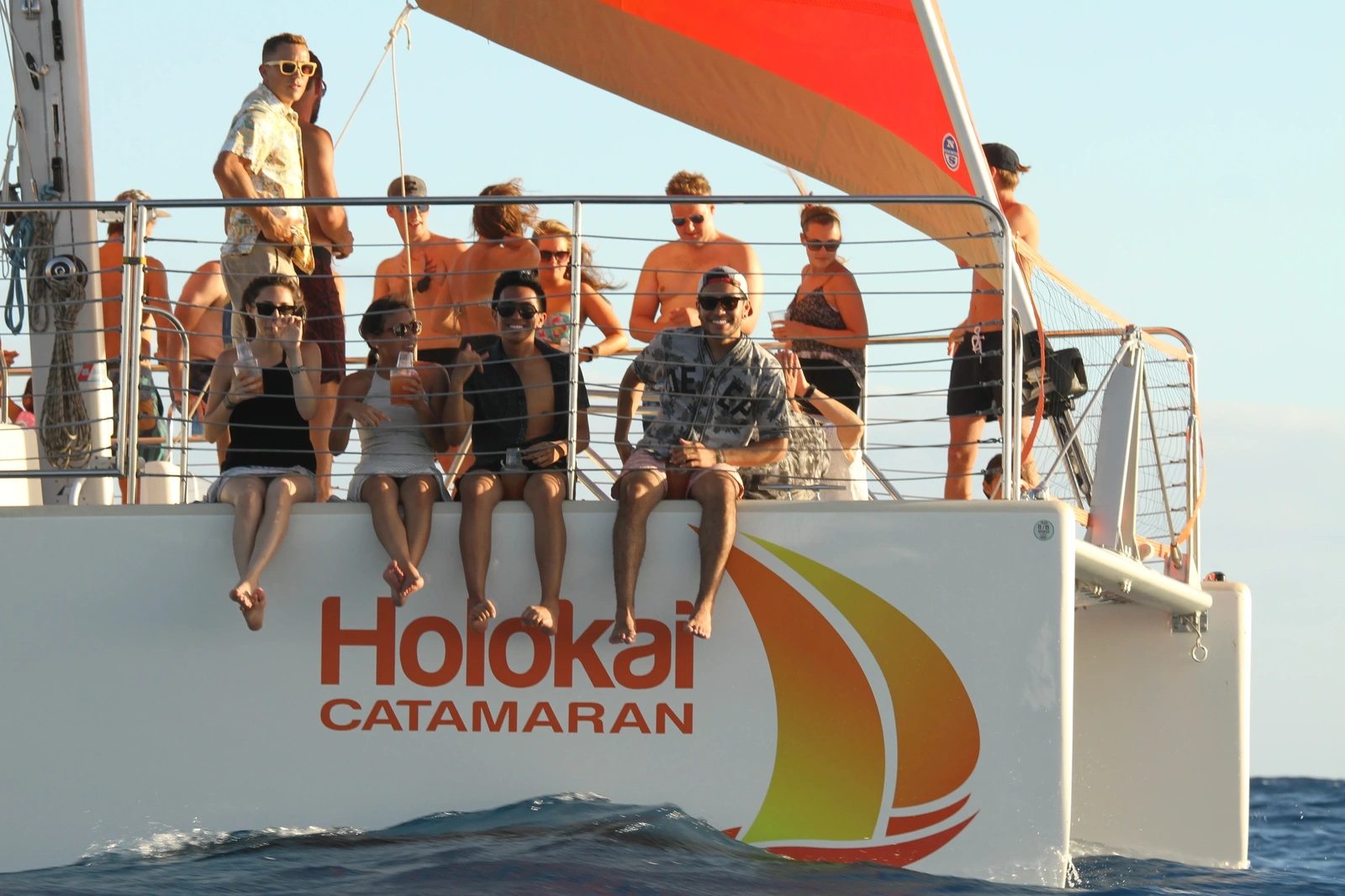 Guests on the netted bow of the catamaran with drinks as the sun sets over the ocean off Waikiki